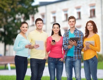 friendship, technology, education, school and people concept - group of smiling teenagers with smartphones and tablet pc computers over campus background