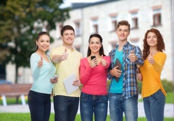 friendship, technology, education, school and people concept - group of smiling teenagers with smartphones and tablet pc computers showing thumbs up over campus background