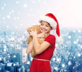holidays, presents, christmas, childhood and people concept - smiling girl in santa helper hat with teddy bear over snowy city background