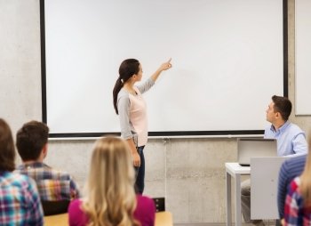 education, high school, technology and people concept - smiling student girl standing in front of white board and teacher with laptop computer in classroom