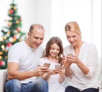 family,  holidays, technology and people - smiling mother, father and little girl with smartphones over living room and christmas tree background
