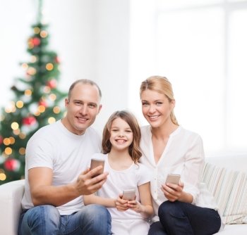 family,  holidays, technology and people - smiling mother, father and little girl with smartphones over living room and christmas tree background