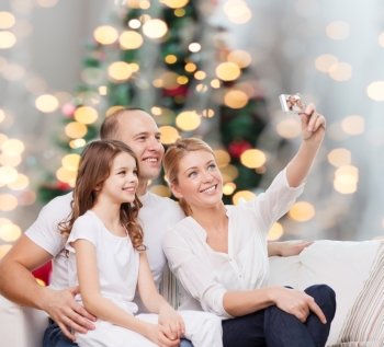 family, holidays, technology and people concept - smiling mother, father and little girl making selfie with camera over christmas tree lights background