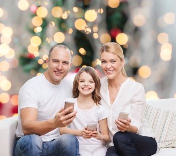 family, holidays, technology and people concept - smiling mother, father and little girl with smartphones over living room and christmas tree background