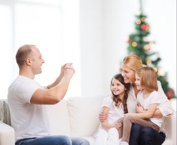 family, holidays, technology and people - smiling mother, father and little girls with camera over living room and christmas tree background