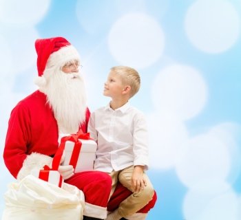 holidays, christmas, childhood and people concept - smiling little boy with santa claus and gifts over blue lights background