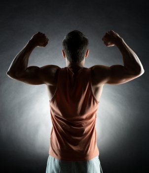 sport, bodybuilding, strength and people concept - young man showing biceps over black background from back