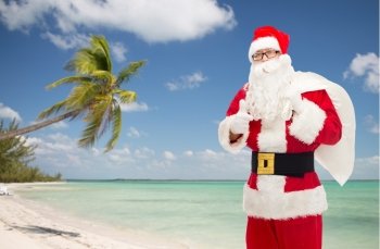 christmas, holidays, gesture, travel and people concept - man in costume of santa claus with bag showing thumbs up over tropical beach background