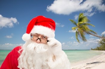 christmas, holidays, travel and people concept - close up of santa claus in glasses winking over tropical beach background