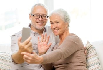 family, technology, age, gesture and people concept - happy senior couple with smartphone making selfie and waving hand at home