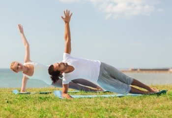 fitness, sport, friendship and lifestyle concept - smiling couple making yoga exercises on mats outdoors
