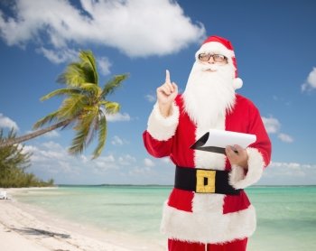 christmas, holidays, gesture, travel and people concept - man in costume of santa claus with notepad pointing finger up over tropical beach background