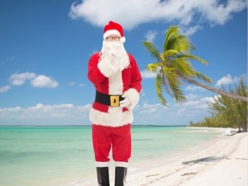 christmas, holidays, travel and people concept - man in costume of santa claus making hush gesture over tropical beach background
