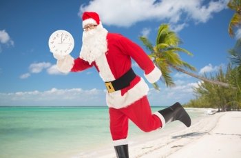 christmas, holidays, travel and people concept - man in costume of santa claus running with clock showing twelve over tropical beach background
