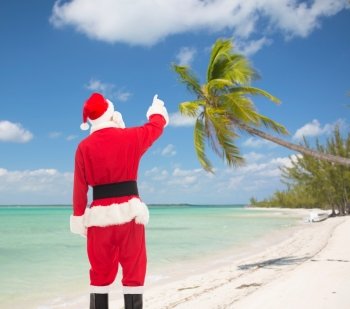 christmas, holidays and people concept - man in costume of santa claus pointing finger from back over tropical beach background