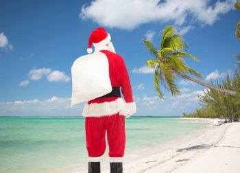 christmas, holidays and people concept - man in costume of santa claus with bag from back over tropical beach background