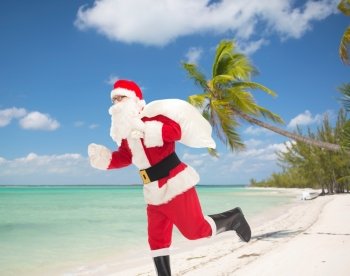 christmas, holidays and people concept - man in costume of santa claus running with bag over tropical beach background