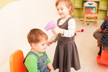 The children are playing a hairdresser game. The girl is holding a hairdryer and making a hairstyle for the boy. The little hairdresser