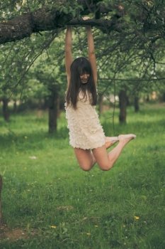 Pretty girl playfully hangs on a tree branch with bent legs. Girl clings to a tree branch