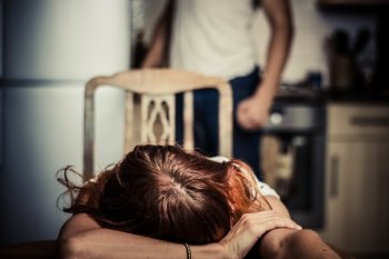 Crying woman in kitchen with abusive partner in the background