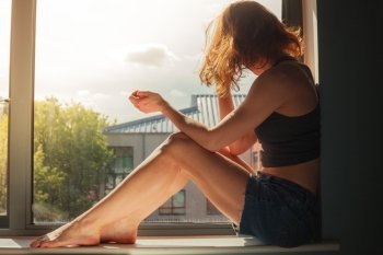 A young woman is sitting on the window sill and looking out on a sunny day