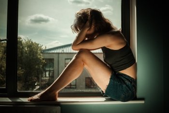 Sad and upset woman is sitting on the window sill