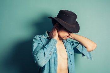 A sexy young cowboy is standing by a blue wall with his shirt unbuttoned