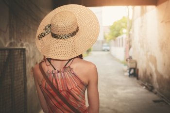 A young woman wearing a hat and a summer dress is standing in an alley at sunset