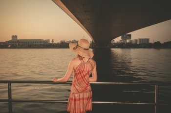 A young woman wearing a hat and summer dress is standing under a bridge at sunset