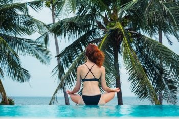 A young woman is sitting in a meditating pose by a swimming pool with palm trees and the ocean in the background