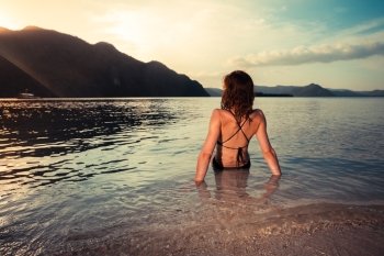A young woman in a bikini is sitting on a tropical beach at sunset