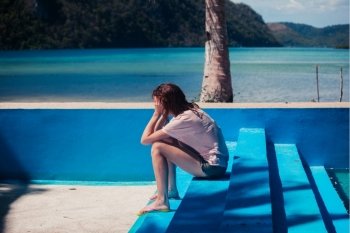 A sad young woman with her head in her hands is sitting in an empty swimming pool by the beach