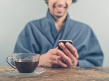 A young man wearing a blue bathrobe is sitting at a table and is using a smart phone while having a cup of coffee in the morning