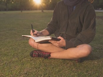 A young woman is sitting on the grass in a park at sunset and is writing in a notebook