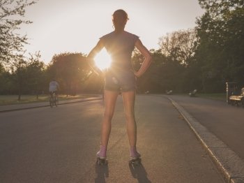 An attractive young woman wearing roller skates is standing in a park admiring the sunset
