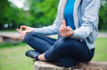 A young woman is sitting in a meditation pose on a tree trunk in the park and is displaying a rude gesture wth her middle finger