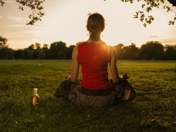 A young woman is meditating in a park at sunset, there is a bottle of health drink next to her