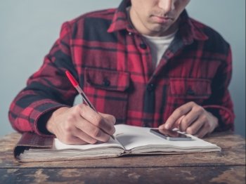 A young man wearing a checkered jacket is using a smartphone and is taking notes in a notebook at a desk