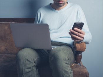 A young man is sitting on a sofa and is using a laptop computer and a smart phone