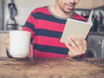 A young man is reading a book and drinking coffee in a kitchen