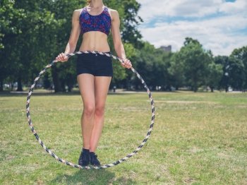 A fit young woman is standing on the grass in a park on a sunny summer day and is holding a hula hoop