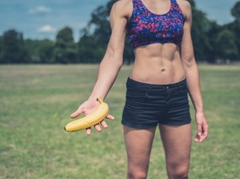 A fit young woman is standing in a park and is offering a banana
