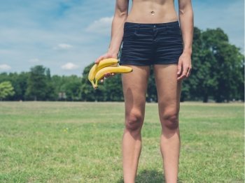 A fit young woman is standing on the grass in a park with two bananas in her hand