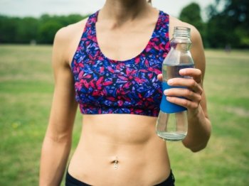 A fit and athletic young woman is standing on the grass in a park with a water bottle in her hand