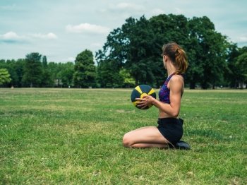 A fit and athletic young woman is sitting on the grass in a park and holding a medicine ball