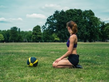 A fit and athletic young woman is sitting on the grass in a park with a medicine ball in front of her