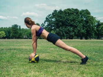 A fit and athletic young woman is doing a plank exercise on a  medicine ball in the park on a sunny summer day