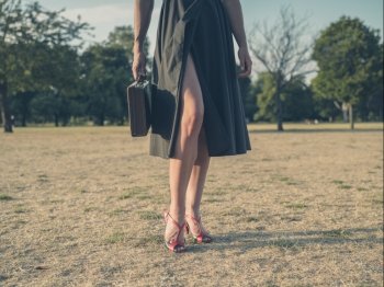 Vintage filtered shot of an elegant young woman wearing a dress and high heels standing in a park with a briefcase