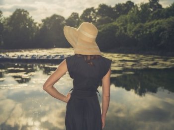Vintage filtered shot of an elegant woman wearing a hat standing by a lake in a park at sunset