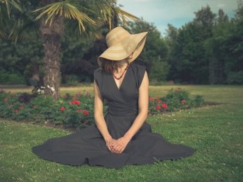Vintage filtered shot of an elegant woman with a hat sitting on the grass in a park by a flowerbed